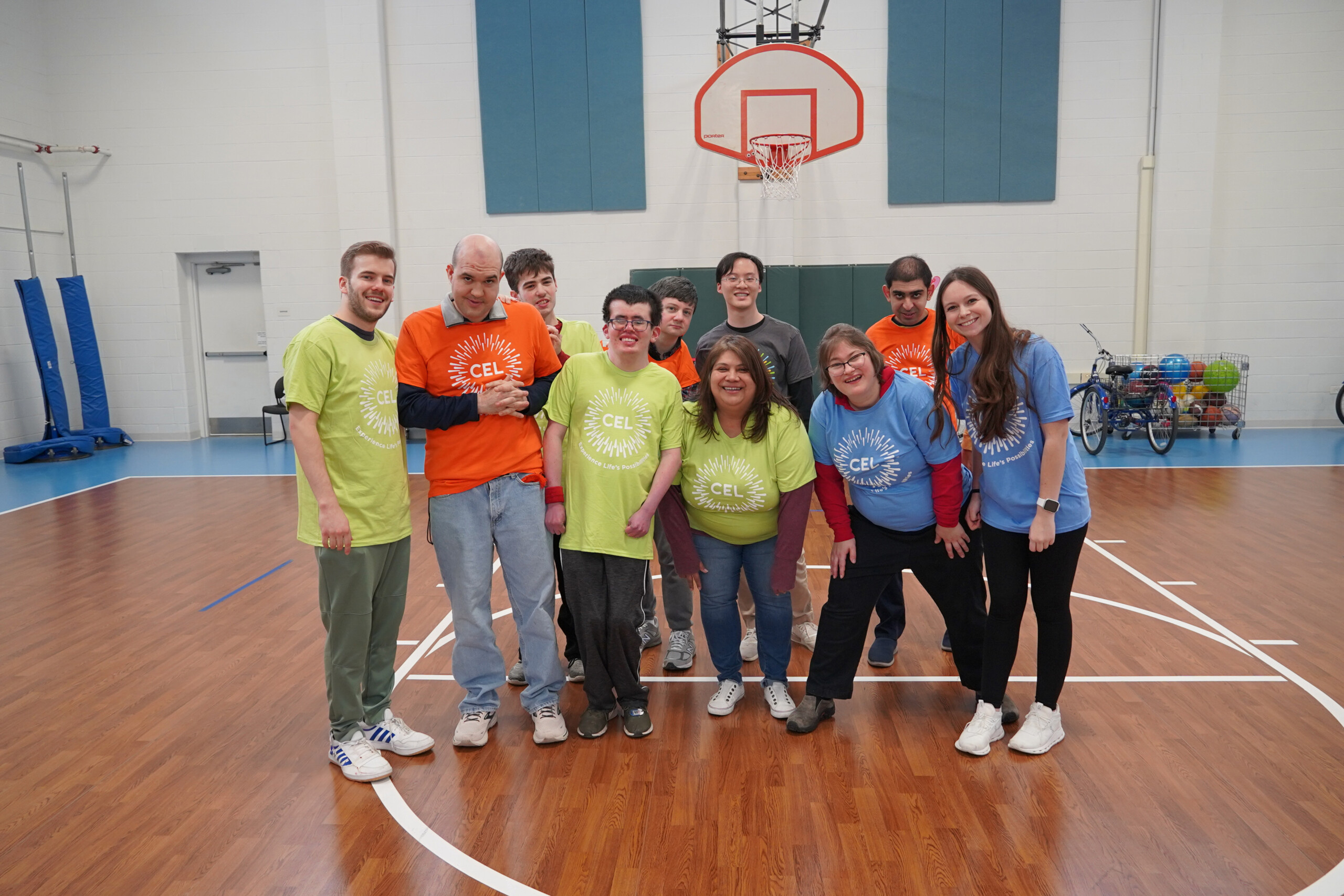 Group of Catalyst life skills program members with staff smiling and wearing different CEL merch in green, blue, and orange, posing in the gym.