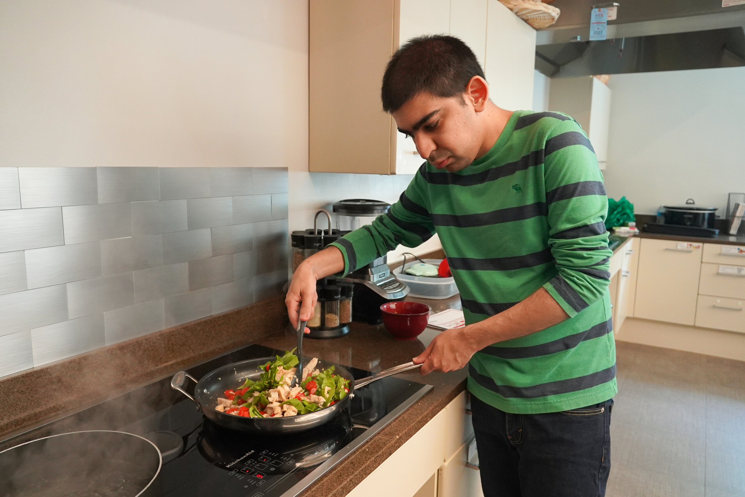 Catalyst member wearing a green and black striped shirt preparing chicken with sliced green and red peppers on a stovetop.