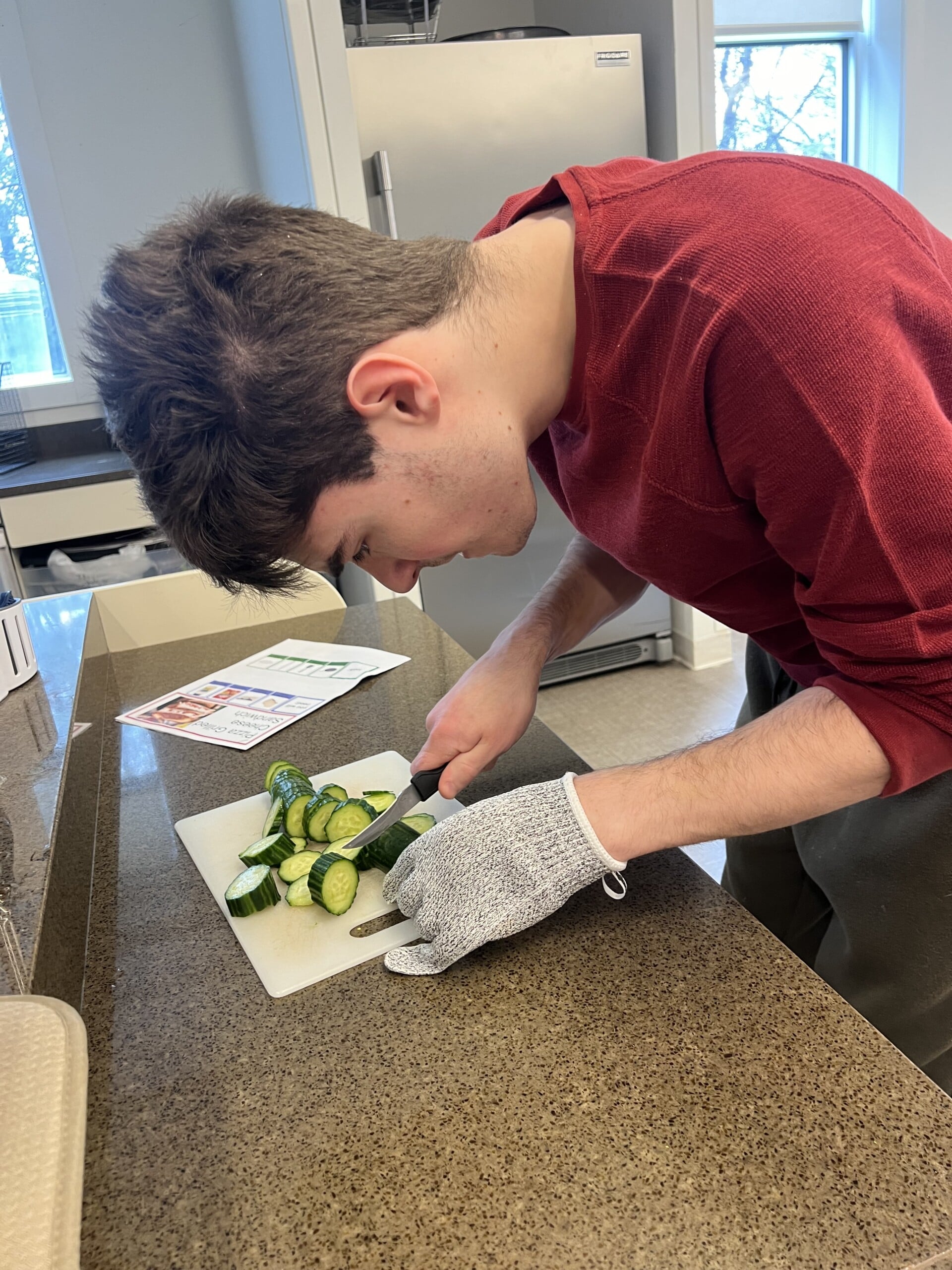 Catalyst member Duncan C cutting cucumbers for a lunch recipe. He is wearing a red three-quarter length sleeve shirt and a cutting glove on his left hand while holding a knife. His other hand holds the cucumber as he focuses on making precise cuts.