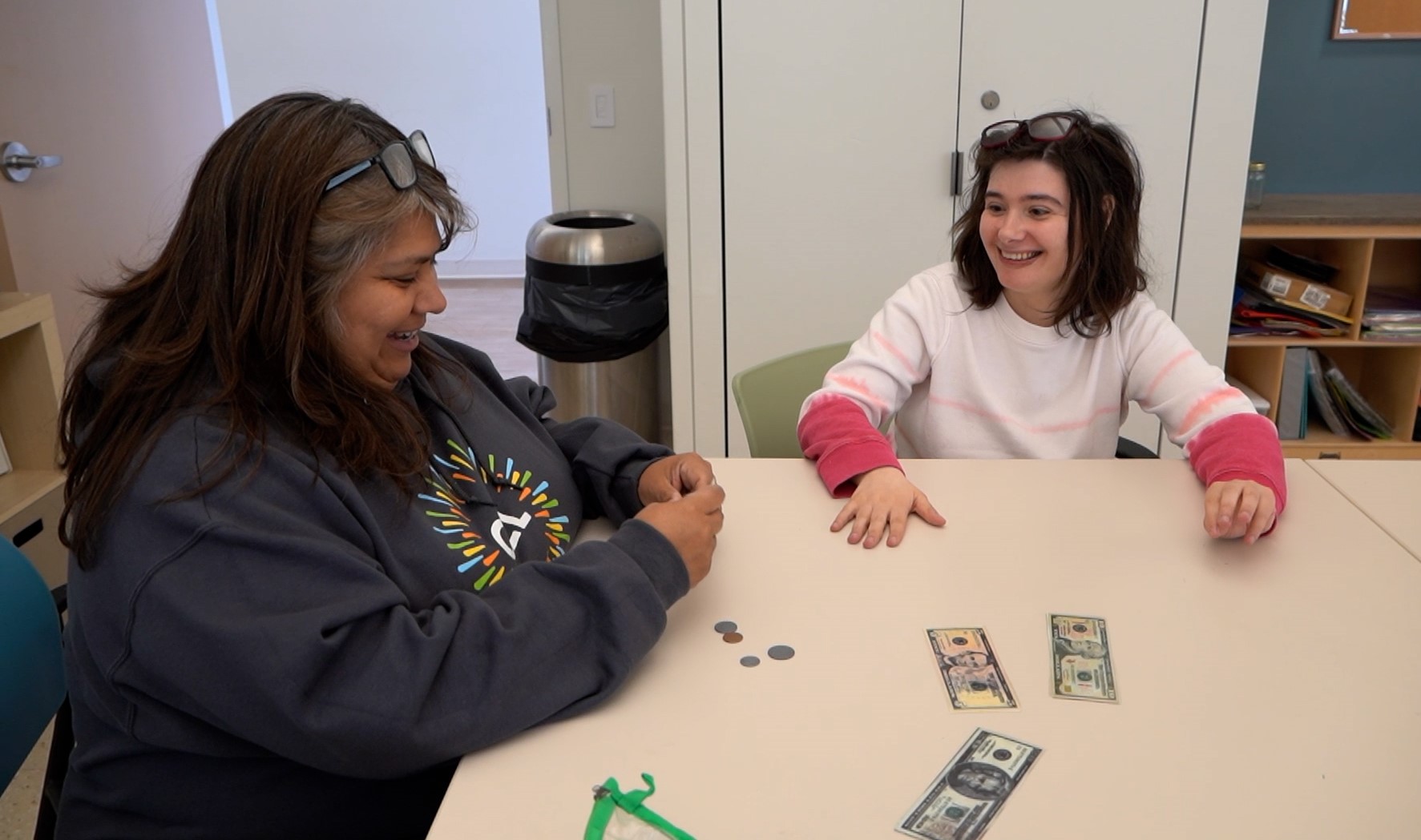 Catalyst member and staff sitting at a table, learning about budgeting with smiles. On the table, there is money and various coins. The staff member is wearing a CEL hoodie, and the member is wearing a pink long sleeve shirt with a white short sleeve shirt over it.