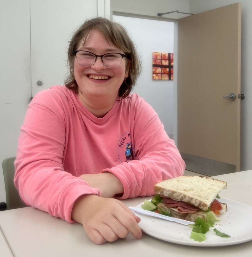 Catalyst member Jenny, wearing a pink long sleeve, sitting at a table and smiling with a sandwich she prepared during meal prep.