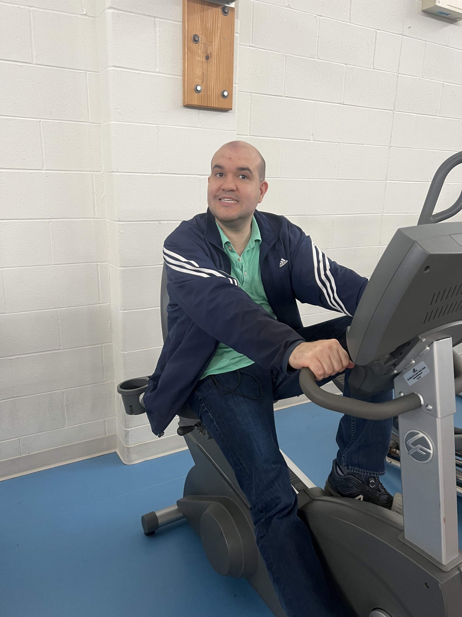 Catalyst life skills program member smiling while working out on a workout bike in the CEL gym. The member is wearing a blue and grey jacket with a green short-sleeve shirt.
