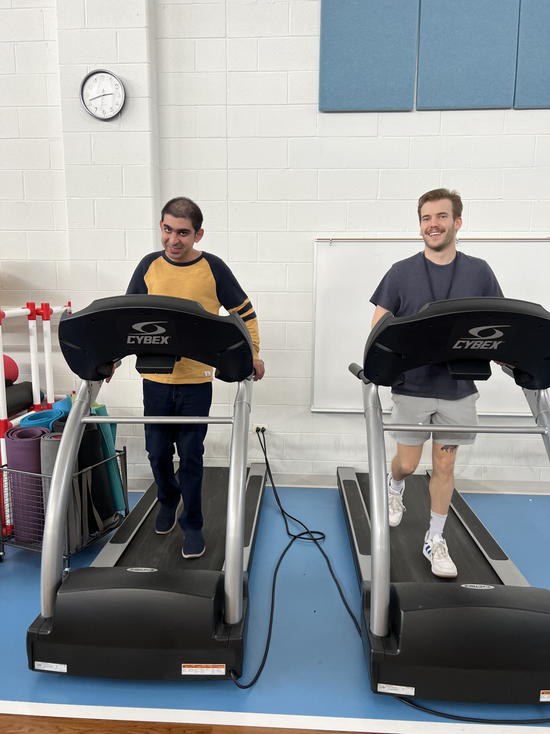 Catalyst member Sam, wearing a black and yellow long sleeve, walking on a treadmill with a staff member in the CEL gym during a health and wellness session.